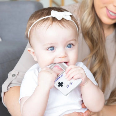 A baby with a white hairband and blue eyes is playfully biting a small, retro-style gaming device. They are sitting in the lap of an adult, who is partially visible and smiling in the background.