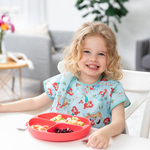 Curly-haired child smiles, wearing a Bumkins Junior Bib: Ariel, eating fruits and veggies from a sectioned plate at the table.