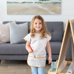 A young girl with curly hair stands in a living room, holding a paintbrush by a wooden easel. She wears Bumkins Short-Sleeved Smock: Wander. A gray sofa with cushions and a landscape painting are in the background.