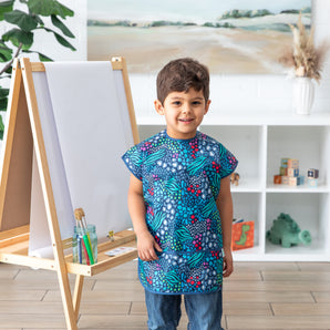 A child in a Bumkins Animal Print smock grins at an easel, their painting shining amid shelves of toys.