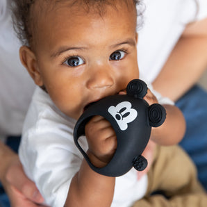 A curly-haired baby chews on a Bumkins black Mickey Mouse teether, made from LFGB-grade silicone, while sitting on an adults lap.