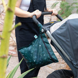 A person in black overalls pushes a stroller on a stone path. Hanging from the handle is a Bumkins Wet Bag: Starry Cactus with celestial, Starry Night prints, set against lush green plants.