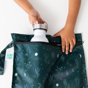 A child’s hands place a white water bottle into a Bumkins Starry Cactus wet bag featuring cacti and mountains on dark green, made from waterproof fabric, set against a white surface.