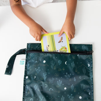 A child puts a yellow-covered book into a dark green Bumkins Wet Bag: Starry Cactus. The waterproof fabric features a cacti and Starry Night Print and rests on a white surface.