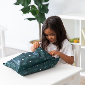 A child with long hair, in a white shirt, opens a Bumkins dark green Wet Bag featuring a Starry Cactus print at the table. A potted plant and shelf provide charm to the cozy setting.