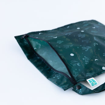 The Bumkins Wet Bag: Starry Cactus is a green reusable bag featuring a cactus and desert-themed pattern. It lies partially open on a white surface, displaying its waterproof fabric, visible zipper, and side label.