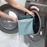 A person places a teal reusable wet bag with a zipper, the Starry Cactus by Bumkins, into an open front-loading washing machine. In the background, theres a white laundry basket with holes, and the countertop is made of white marble.