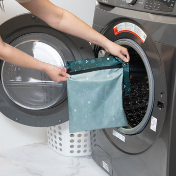 A person holds a Bumkins Wet Bag: Starry Cactus in front of a front-loading washer on a marble floor next to a white laundry basket.