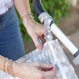 A close-up shows someone attaching the Bumkins Wet Bag: Lunar Phase, featuring moon phases and heat-sealed seams, to a black and silver stroller handle. The persons arms, adorned with bracelets, are visible against a backdrop of blurred green foliage.