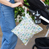 A person in jeans holds a stroller where a Bumkins Wet Bag: Cacti hangs from the handle. In the cup holder, theres a cup with a straw, set against lush greenery.