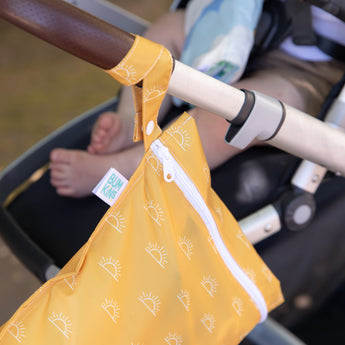 The Bumkins Wet Bag: Sunshine is clipped to a stroller with baby legs visible, featuring sun patterns and a smooth zipper closure.