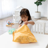 A young girl at a white table opens Bumkins Wet Bag: Sunshine, with a playhouse and toys brightening the airy room.