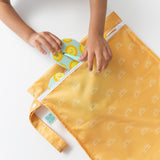 A childs hands tuck a colorful diaper into the Bumkins Wet Bag: Sunshine on a white surface.