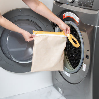 A person holds a Bumkins Wet Bag: Sunshine in front of a gray front-load washing machine.