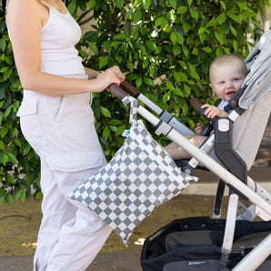 Person in white outfit pushes a stroller with Bumkins Charcoal Check Wet/Dry Bag attached. Green foliage in the background.