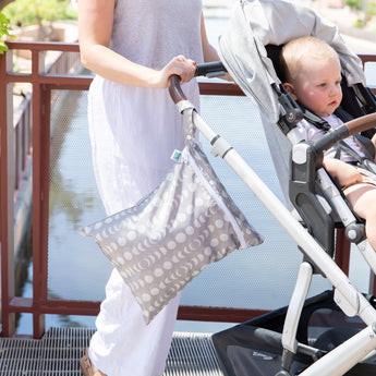 A person wearing white pants and a gray top pushes a baby stroller on a bridge. A child sits in the stroller, looking to the side, while a Bumkins Wet/Dry Bag: Lunar Phase is attached to the handle, ideal for any outing with its waterproof fabric.