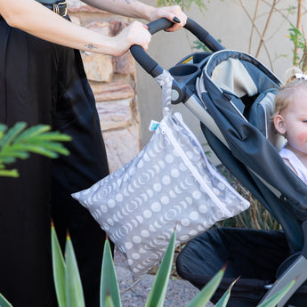 A person pushes a stroller with a small child while a Bumkins Wet/Dry Bag: Lunar Phase, made of waterproof fabric with a moon phase pattern, hangs from the handle. Green plants and a stone wall are in the background.