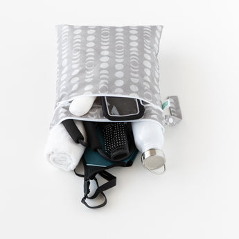 The open Bumkins Wet/Dry Bag: Lunar Phase, featuring a stylish gray and white pattern crafted from waterproof fabric, reveals a smartphone, towel, hairbrush, stainless steel water bottle, and earphones spilling onto the white surface.