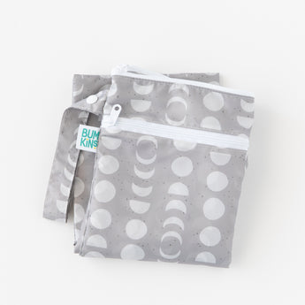 The Bumkins Wet/Dry Bag: Lunar Phase is gray with white crescent and circle patterns, made from waterproof fabric. It has a zipper closure, small handle, and a visible BUMKINS tag. The bag is neatly folded for convenience.