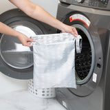 A customer places the Bumkins Wet/Dry Bag: Lunar Phase, featuring a subtle polka dot pattern on waterproof fabric, into a front-loading washing machine. In the backdrop are a laundry basket and marble countertop.