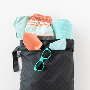 A Rich Black Wet/Dry Bag by Bumkins with sunglasses, towel, swimwear, and toy shovels peeks atop a white surface.