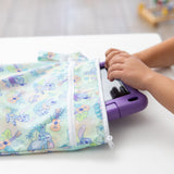 Childs hands play on a purple toy piano beside a colorful Bumkins Wet/Dry Bag: Aloha Stitch on a white table.