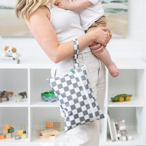 A woman holds a child and a Bumkins Wet/Dry Clutch: Charcoal Check in a toy-filled room.