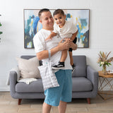 In a living room with a gray sofa, colorful abstract art, and decorative plants on wooden flooring, a smiling man in a white shirt holds a young boy. Hes carrying Bumkins stylish Wet/Dry Clutch: Lunar Phase, crafted from waterproof fabric.