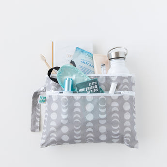 The Bumkins Wet/Dry Clutch: Lunar Phase in gray, featuring a lunar phases pattern, holds a water bottle, book, sheet mask, toothbrush, deodorant, and teeth whitening strips. It includes two visible compartments and is presented against a plain white background.