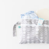 The Bumkins Wet/Dry Clutch: Lunar Phase is a gray diaper bag with a crescent moon pattern that opens to reveal blue diapers and diaper rash cream inside. Made from waterproof fabric, it features a zipper and strap, showcased on a white background.