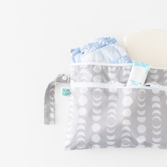 The Bumkins Wet/Dry Clutch: Lunar Phase is a gray diaper bag with a crescent moon pattern that opens to reveal blue diapers and diaper rash cream inside. Made from waterproof fabric, it features a zipper and strap, showcased on a white background.