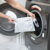 A Bumkins Wet/Dry Clutch: Lunar Phase in gray is placed into a front-loading washing machine, while a nearby white laundry basket suggests an organized routine.