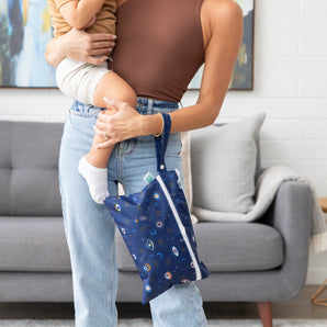 Woman holds child in living room, carrying a Bumkins Wet/Dry Clutch: Evil Eye. Sofa and abstract art accentuate the cozy scene.