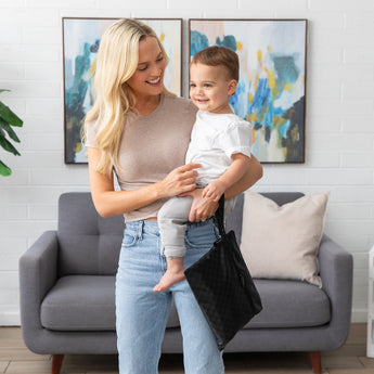 A woman holding a toddler smiles in a living room with abstract art and a gray sofa, a Bumkins Wet/Dry Clutch: Rich Black on her wrist.