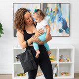 A woman in workout clothes holds a smiling toddler and a Bumkins Wet/Dry Clutch: Rich Black in a colorful room with abstract art.