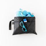 A Bumkins Wet/Dry Clutch in Rich Black with blue sunglasses, a towel, shoes, a bottle, and sunscreen against a white background.