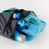 The Rich Black Wet/Dry Clutch by Bumkins lays open on a white background, holding a blue bottle, shoes, towel, swimsuit, and sunglasses.