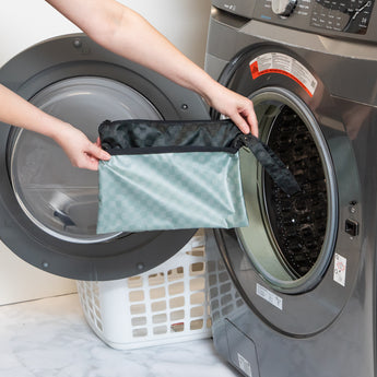 A person places a Bumkins Wet/Dry Clutch: Rich Black into a washing machine next to a white laundry basket.