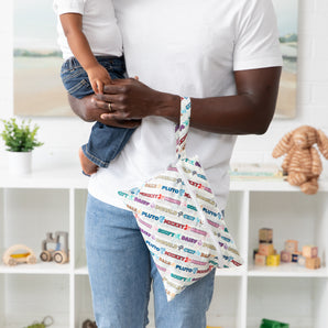 A man holds a child and a Bumkins Wet/Dry Clutch: Mickey & Friends Varsity on his arm; toys and plant on shelves behind them.