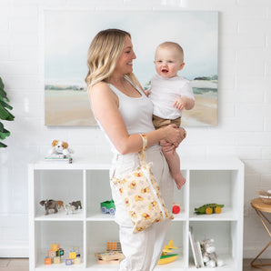 A woman, wearing a white top and pants, stands in a bright room, holding a smiling baby and a Bumkins Wet/Dry Clutch: Winnie Loves Hunny.