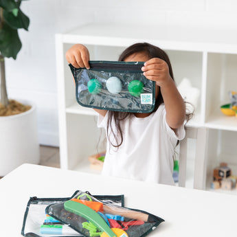 A child in a white shirt displays a Bumkins Clear Travel Bag 3 Pack: Starry Cactus, filled with green and white items, while another reusable bag with colorful objects rests on the table. A plant and toy-adorned shelves enhance the backgrounds charm.