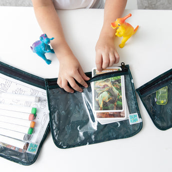 A child places a dinosaur card into a Bumkins Clear Travel Bag from the Starry Cactus 3 Pack on a white table. Two colorful dinosaur toys are nearby, while other reusable bags contain markers and additional cards.