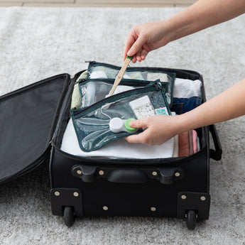 A person is packing a suitcase filled with clothes and a TSA-compliant Clear Travel Bag 3 Pack: Starry Cactus by Bumkins for personal care items while holding a wooden toothbrush.