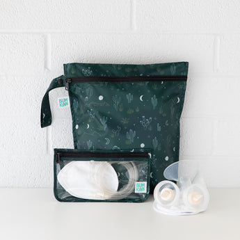 A BUMKINS dark green wet bag featuring a cactus and moon design stands upright. In front, a clear bag, part of the Clear Travel Bag 3 Pack: Starry Cactus, holds breast pump parts like tubing and silicone flanges. These reusable TSA-compliant bags sit on a white surface for easy organization and transport.