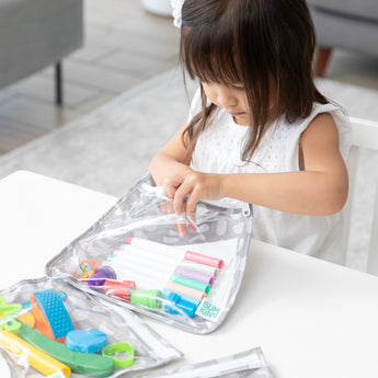 A child in a white dress opens a Bumkins Clear Travel Bag 3 Pack: Lunar Phase  filled with colorful markers and scissors at a white table.