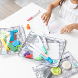 A child draws with colorful markers at a white table, surrounded by vibrant craft supplies in Bumkins Clear Travel Bag 3 Pack: Lunar Phase.