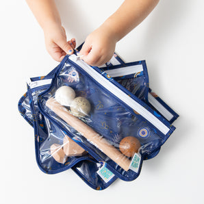 A child unzips a Bumkins Clear Travel Bag 3 Pack: Evil Eye with toys inside on a white table.