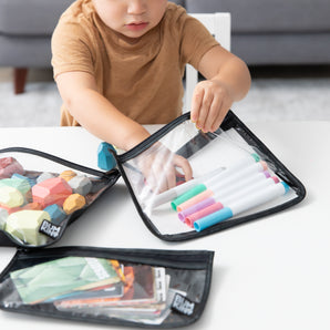 A child organizes markers into a Bumkins Clear Travel Bag at a table, surrounded by reusable bags with geometric shapes and photos.