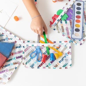 A child grabs art supplies from a Bumkins Clear Travel Bag 3 Pack: Mickey & Friends Varsity on a white table.