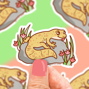 Cartoon gecko vinyl sticker by Turtle Soup: smiling yellow gecko on a rock with pink flowers, surrounded by more colorful geckos.